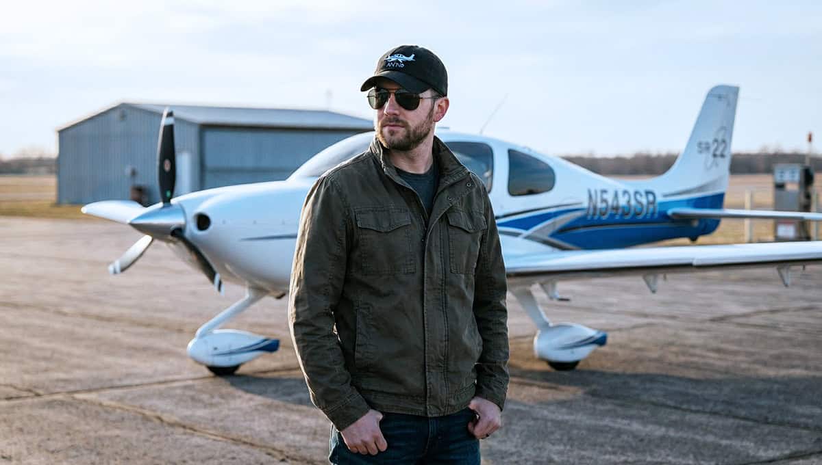 Man in ramp-ready pilot hat standing beside a small private plane outdoors.